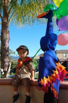 a little boy standing next to a blue bird with balloons on it's head