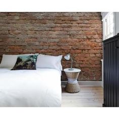 a bed with white sheets and pillows in front of a brick wall, next to a night stand