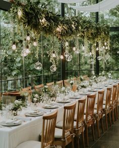 a long table is set with white linens, greenery and hanging glass balls