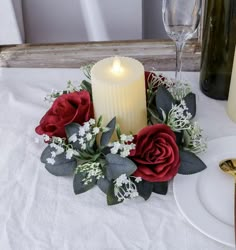 a candle and some red roses on a white table cloth with wine bottles in the background