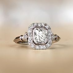 This item has just been reserved by another customer. Please contact us to be notified if it becomes available.  A lovely center antique cushion cut diamond features a bezel set in a handcrafted platinum mounting. The center diamond is approx 1.07 carats, J color, and VS2 clarity, and is surrounded by a halo of diamonds. Diamond-set fleur-de-lis motif shoulders lead to a triple-wire shank. This platinum ring is decorated with an openwork under-gallery. ✦ DIAMOND SPECIFICATIONS:    Center Diamond Silver Cushion Cut Art Deco Ring, Formal Art Deco Cushion Cut Diamond Ring, Art Deco Diamond Jewelry Cushion Cut, Art Deco Platinum Cushion Cut Jewelry, Art Deco Cushion Cut Platinum Jewelry, Antique Cushion Cut Diamond Engagement Ring, Antique Cushion Cut Diamond Ring, Antique Cushion Cut Diamond, Antique Cushion Cut