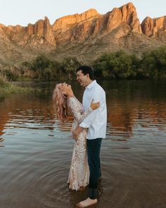 a man and woman are standing in the water near some mountains with their arms around each other