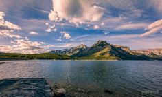 a lake with mountains in the background under a blue sky filled with white fluffy clouds