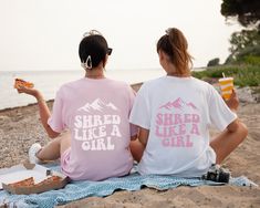 two women sitting on the beach eating pizza and drinking sodas while looking out at the ocean