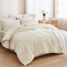 a bed with white comforter and pillows in a room next to a window on a hard wood floor