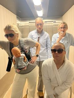 four people standing in an elevator with one holding a doll and the other wearing sunglasses