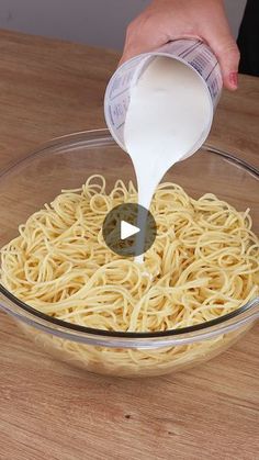 someone pouring milk over noodles in a glass bowl on a wooden counter top with a person's hand