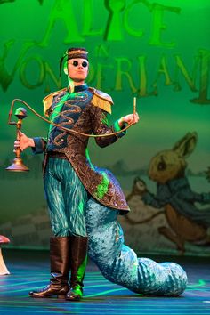 a man dressed as a mermaid on stage with a bell in his hand and an instrument in his other hand