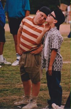 two young boys standing next to each other on a grass covered field with people in the background