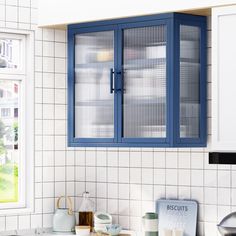 a kitchen with blue cupboards and white tiles on the wall, in front of a window