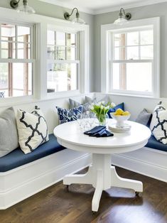 a white table with blue cushions and pillows on it in front of two large windows