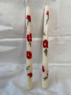 two candles with red flowers on them sitting next to each other in front of a white sheet