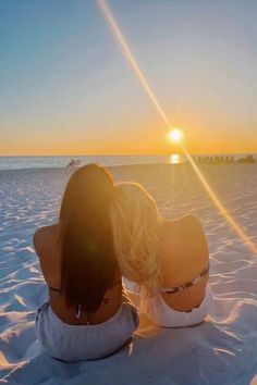 two women are sitting in the sand at sunset, looking out over the water and into the distance