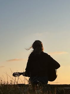 a woman sitting in the grass with her guitar looking at the sun setting behind her