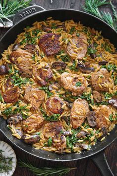 a skillet filled with pasta and meat on top of a wooden table next to fresh herbs