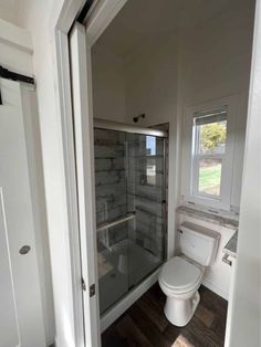 a white toilet sitting next to a shower in a bathroom under an open window on top of a hard wood floor