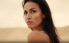 a woman with long black hair and brown eyes looks off into the distance while standing in front of a desert landscape