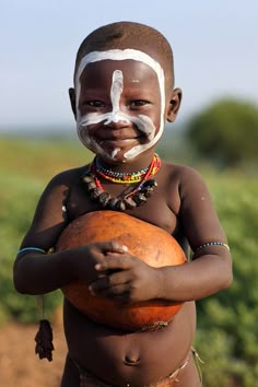 a young boy with white paint on his face and body holding an orange ball in front of him