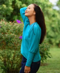 a woman standing in the grass with her hands behind her head and looking up into the sky