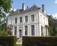 a large white house with two chimneys and blue doors in front of some hedges on a sunny day