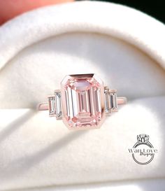 a pink diamond ring sitting on top of a white cloth covered cushioned pillow, with three baguets surrounding it