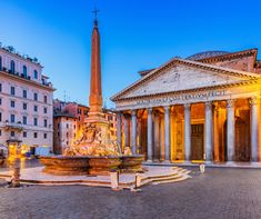 an old building with columns and a fountain in front of it at dusk, surrounded by other buildings