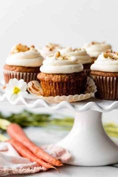 carrot cupcakes with cream cheese frosting on a white cake stand next to fresh carrots