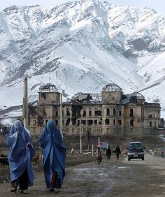 two women in blue robes walking towards a large building with snow covered mountains behind them