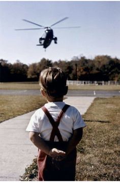 a young boy standing in front of a helicopter flying over the top of a field