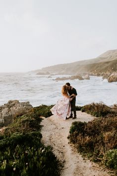 a man and woman embracing each other while standing on a path near the ocean in front of some bushes