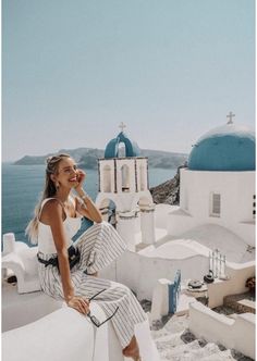 a woman sitting on top of a white building next to the ocean with blue domes