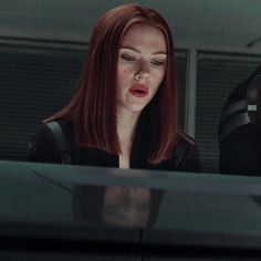 a woman with red hair standing next to a man in a black suit and helmet