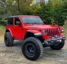 a red jeep parked on top of a dirt field next to trees and bushes in the background