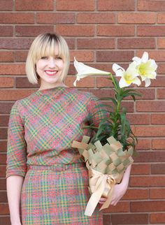 a woman in a dress holding a potted plant next to a brick wall and smiling at the camera