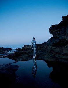 a man is standing in the water with his reflection on the ground and rocks behind him