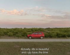 a red car driving down a road next to a lush green field with trees and bushes