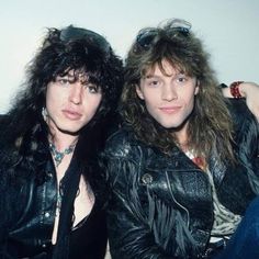 two people sitting next to each other wearing black leather jackets and hair in front of a white wall