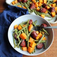 two white bowls filled with green beans, potatoes and sausage on top of a wooden table