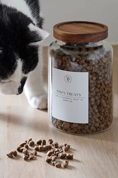 a black and white cat standing next to a glass jar filled with dry food on top of a wooden table