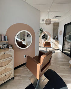 A caramel-colored salon chair stands in front of an LED-mounted mirror. The wall has a light pink arched design and to the left stands a boho-inspired styling station. Minerva Shampoo Bowl, Hair Salon Shampoo Bowls, Salon Station Minerva Beauty, Salon Chairs Minerva Beauty, Shampoo Bowl And Chair, Beauty Bar Ideas