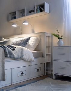 a white bed sitting under a window next to a dresser with drawers and a lamp