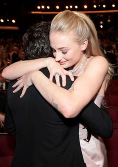 two people hugging each other in front of an audience at a convention or event, one person is wearing a black dress and the other has her arm around her neck