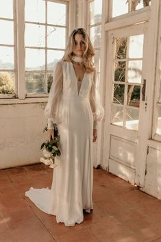 a woman in a white dress is standing by some windows and holding a flower bouquet