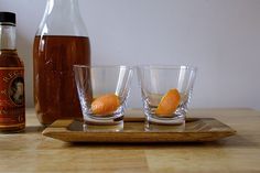 three shot glasses with orange peels sit on a wooden tray next to a bottle of syrup