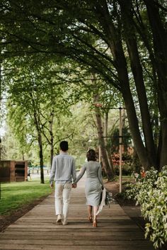 a man and woman walking down a wooden path holding hands in the middle of trees