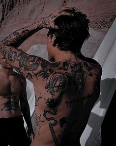 two men standing next to each other with tattoos on their backs and arms, one man has his hands behind his head