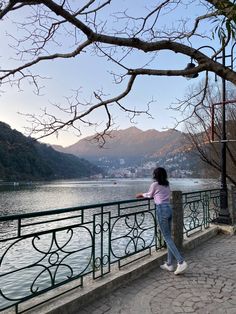 a woman standing on the edge of a bridge next to a body of water with mountains in the background