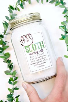 a hand holding a glass jar filled with white stuff and green leaves around it, on top of a table