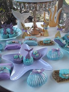 a table topped with plates covered in blue and purple mermaid themed food items on top of a white table cloth