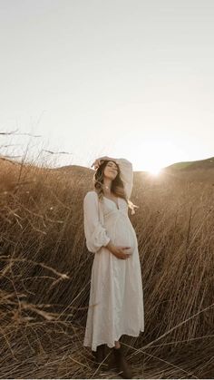 a pregnant woman wearing a white dress and bonnet standing in tall grass with the sun behind her